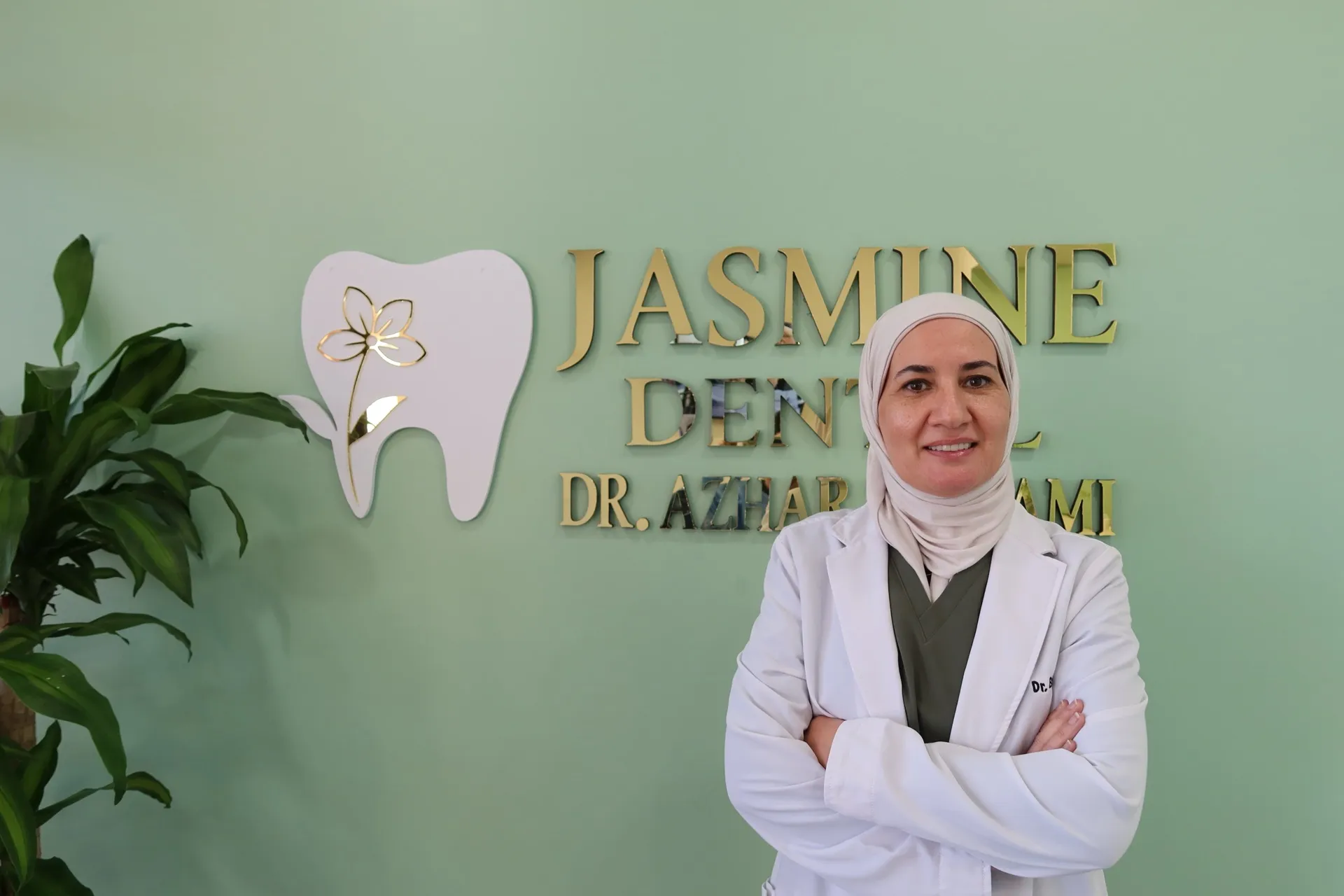 Dr. Azhar Bustami in her white coat at Jasmine Dental in Tampa, standing in front of the practice sign: a jasmine flower set inside a tooth, with her name in gold lettering on the green wall behind her.